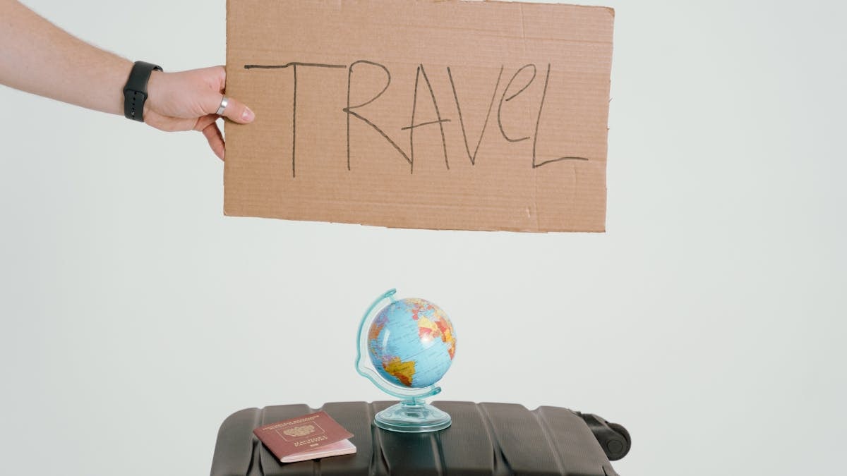 A hand holds a travel sign above a globe and suitcase, symbolizing wanderlust.